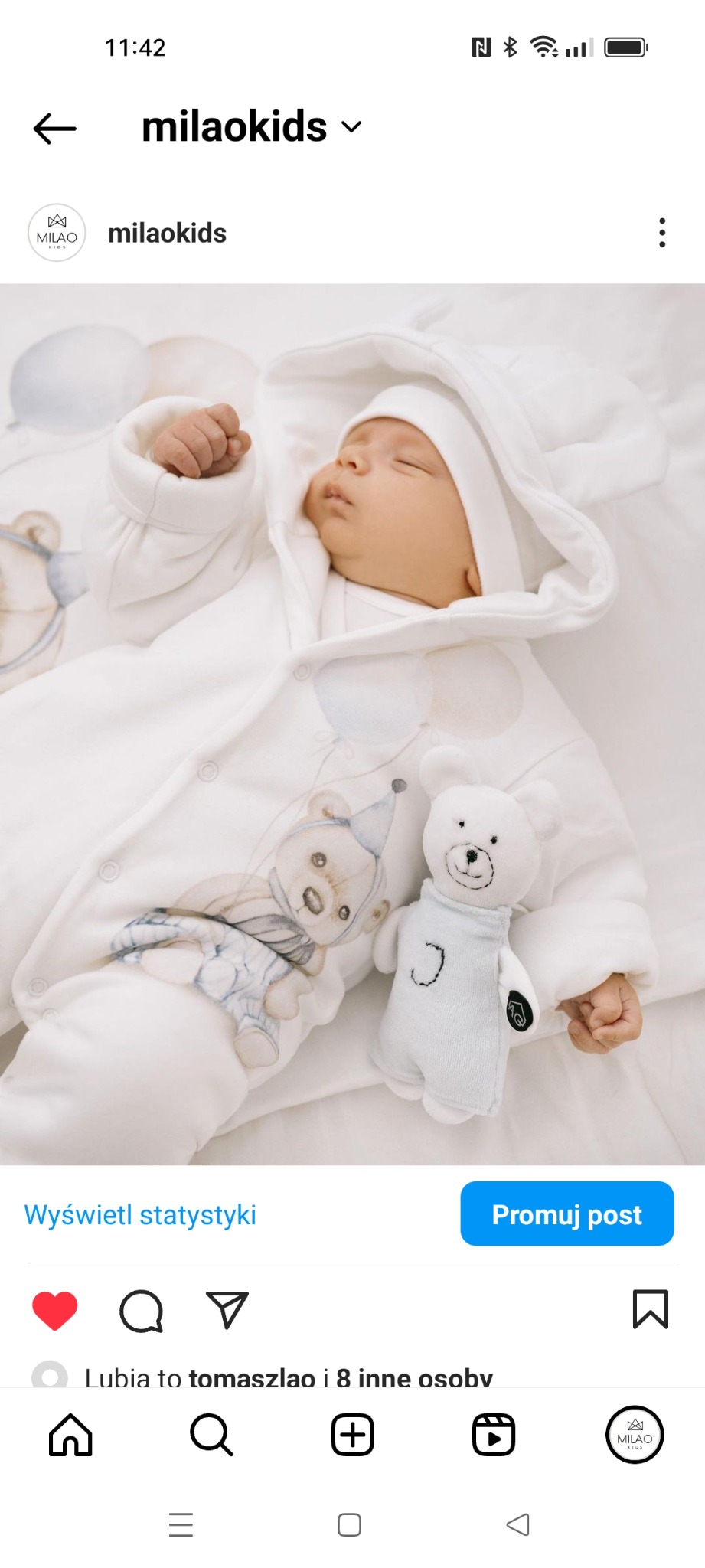 Śpiący niemowlak ubrany w biały kombinezon z kapturem i nadrukiem misia z balonami, obok leży pluszowy miś w błękitnym kombinezonie.