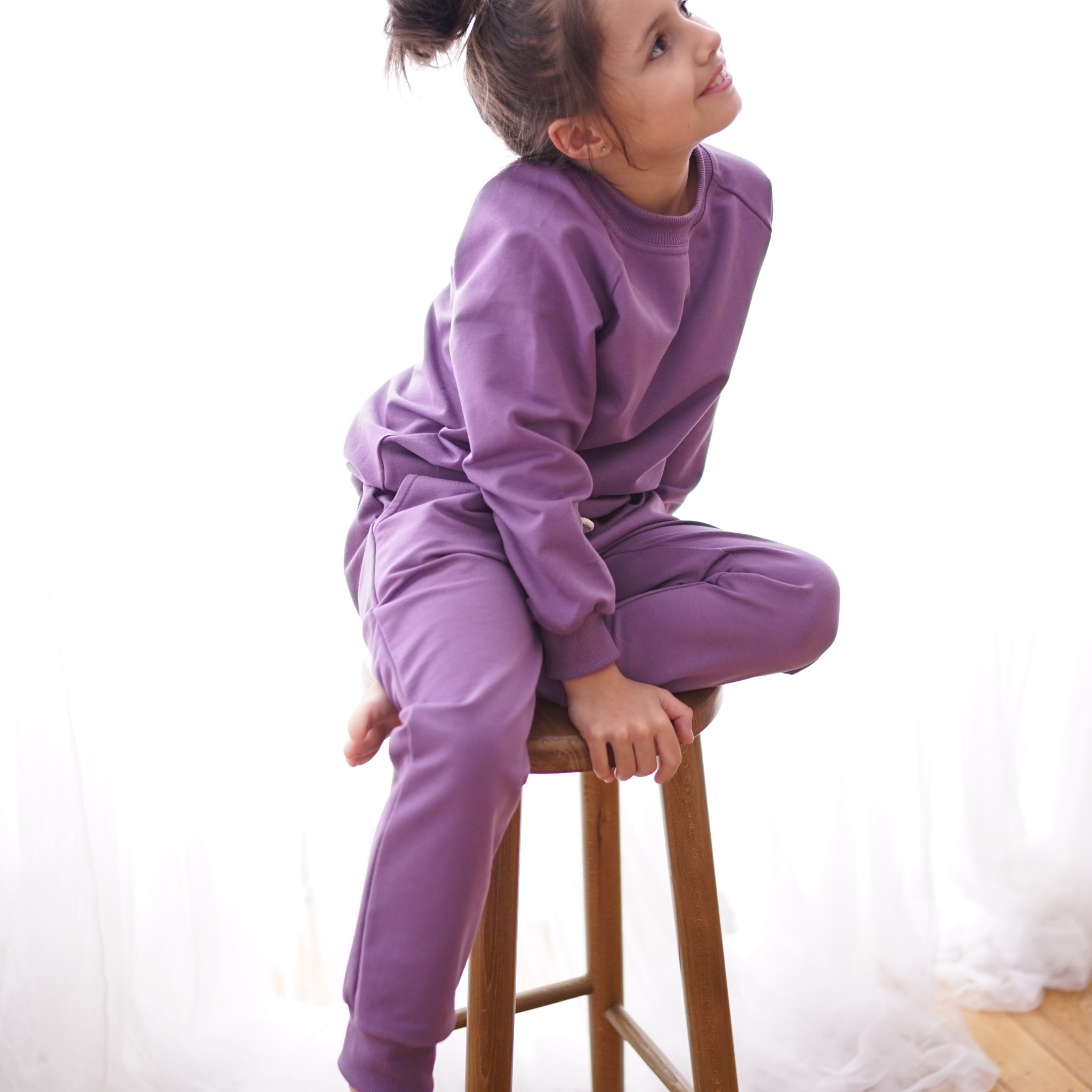 Dziewczynka siedzi na drewnianym stołku, ubrana w fioletowy dres, spoglądając w górę na tle białej zasłony.