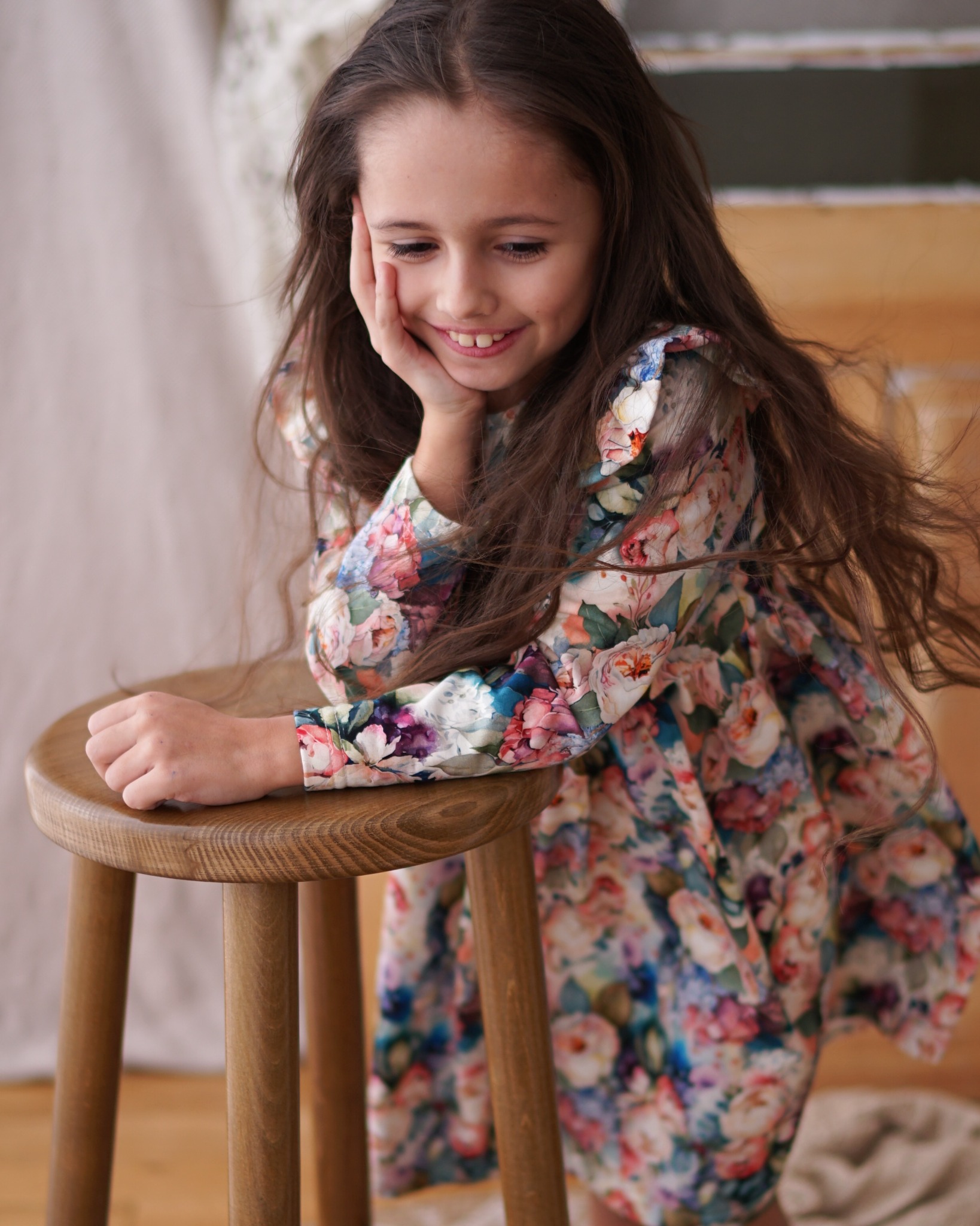 Dziewczynka z długimi brązowymi włosami w sukience w kwiaty, oparta o drewniany taboret, uśmiecha się, patrząc w dół.