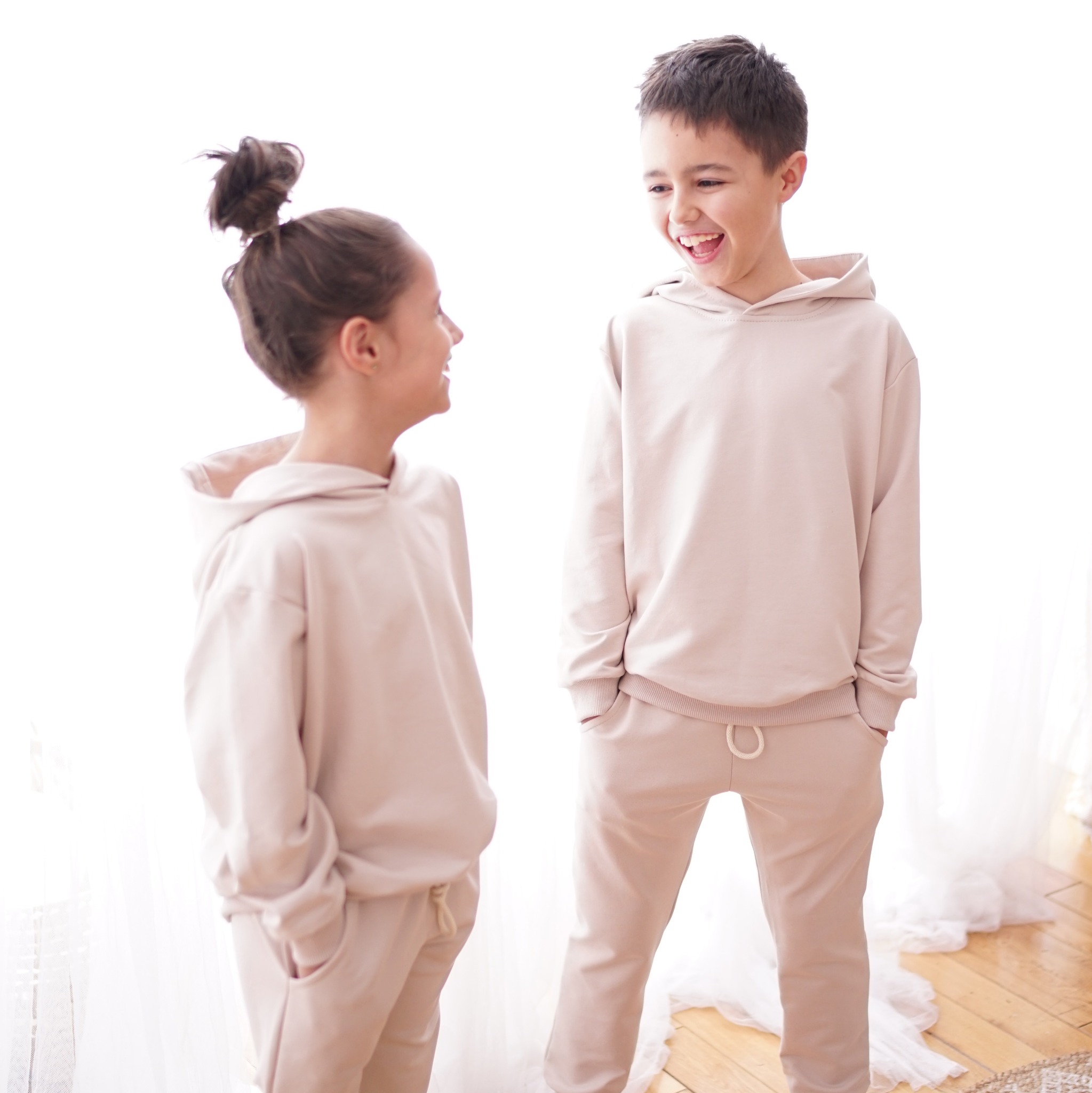 Dwoje dzieci, dziewczynka z włosami upiętymi w kok i chłopiec, ubrani w beżowe, dresowe komplety z kapturem, stoją naprzeciwko siebie i śmieją się na tle jasnej zasłony.