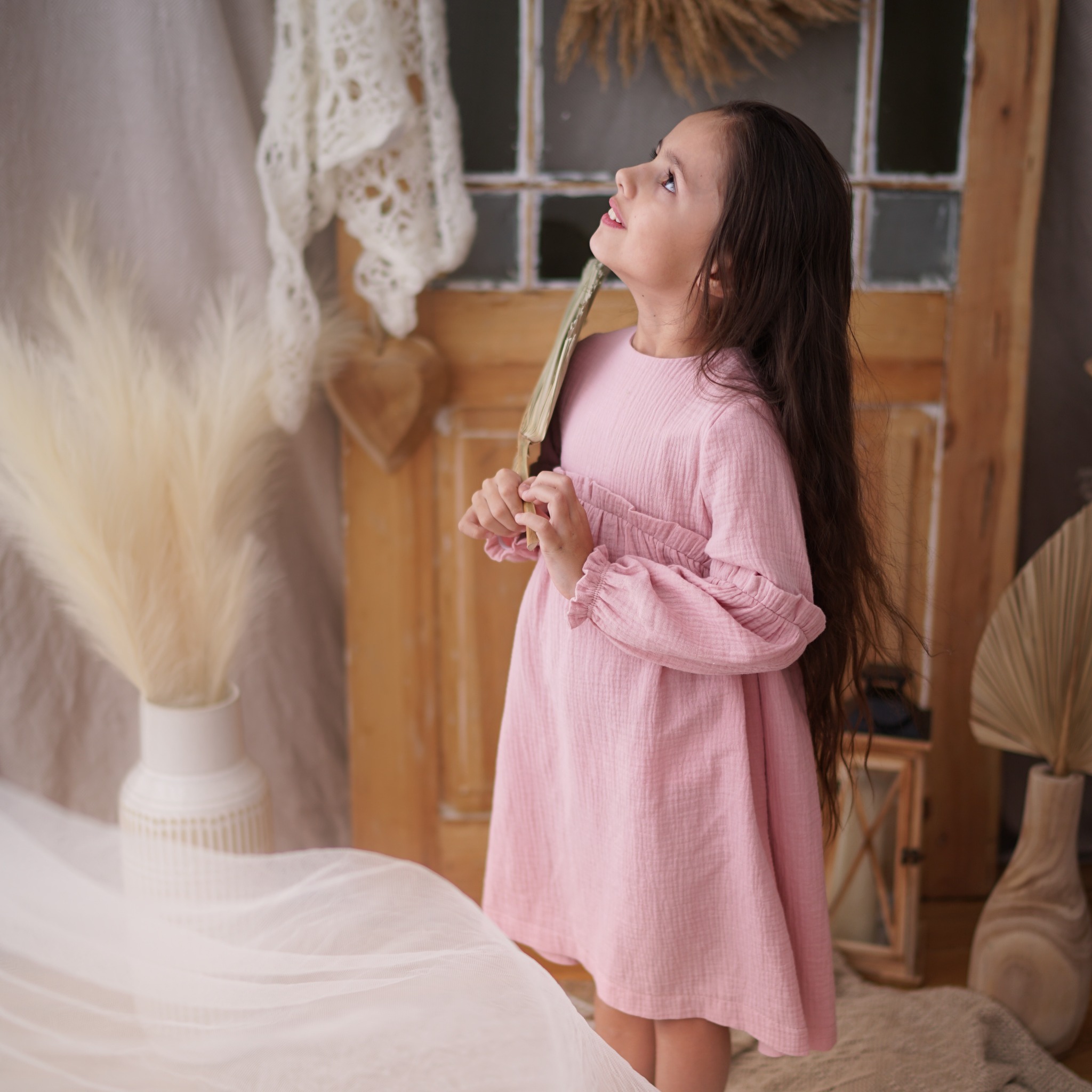 Dziewczynka w różowej, muślinowej sukience z długimi rękawami, trzymająca wachlarz na tle rustykalnych dekoracji i drewnianych drzwi.