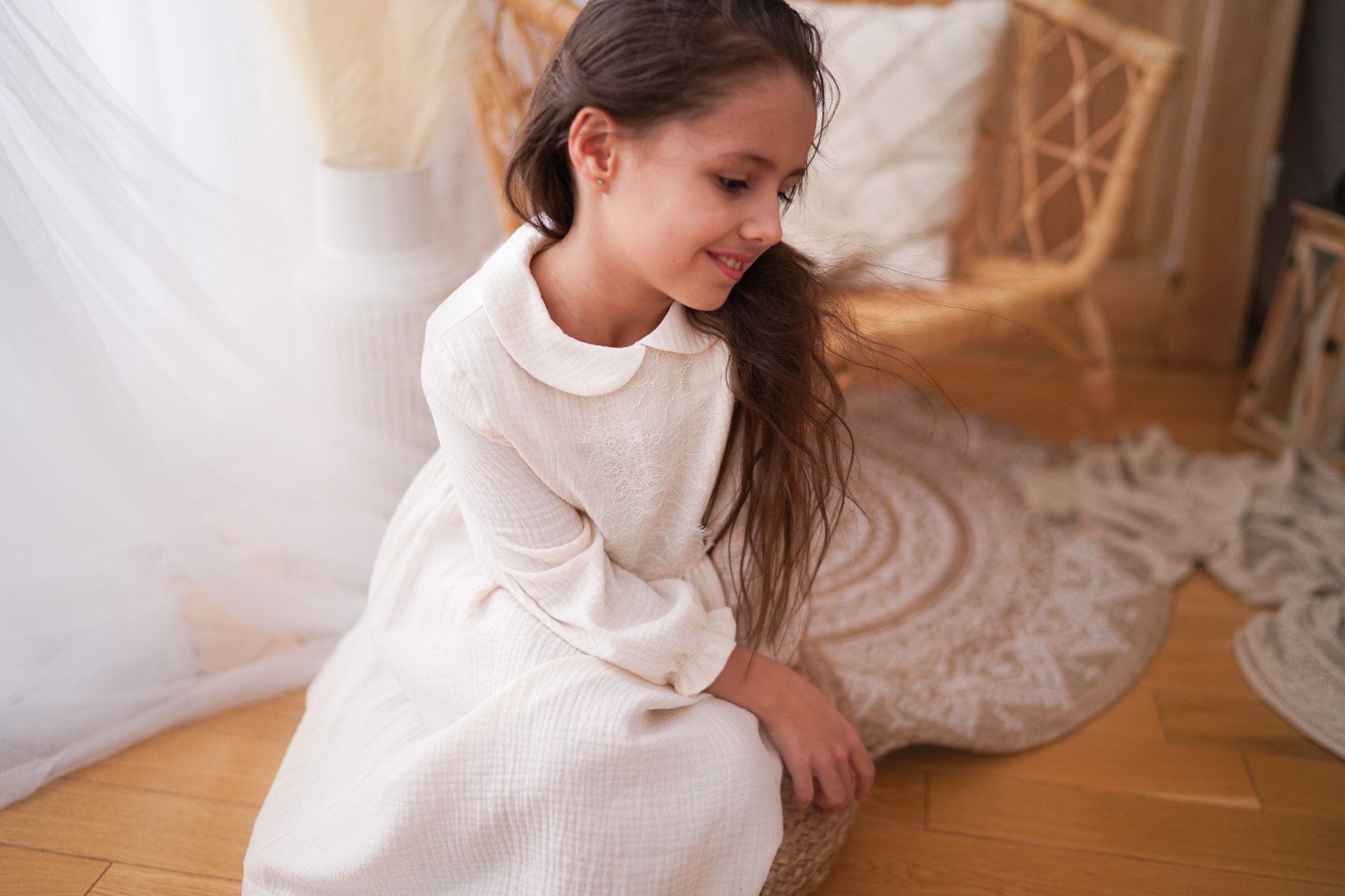 Uśmiechnięta dziewczynka w kremowej, lnianej sukience z kołnierzykiem siedzi na podłodze, w tle rattanowe krzesło i dekoracje w stylu boho.