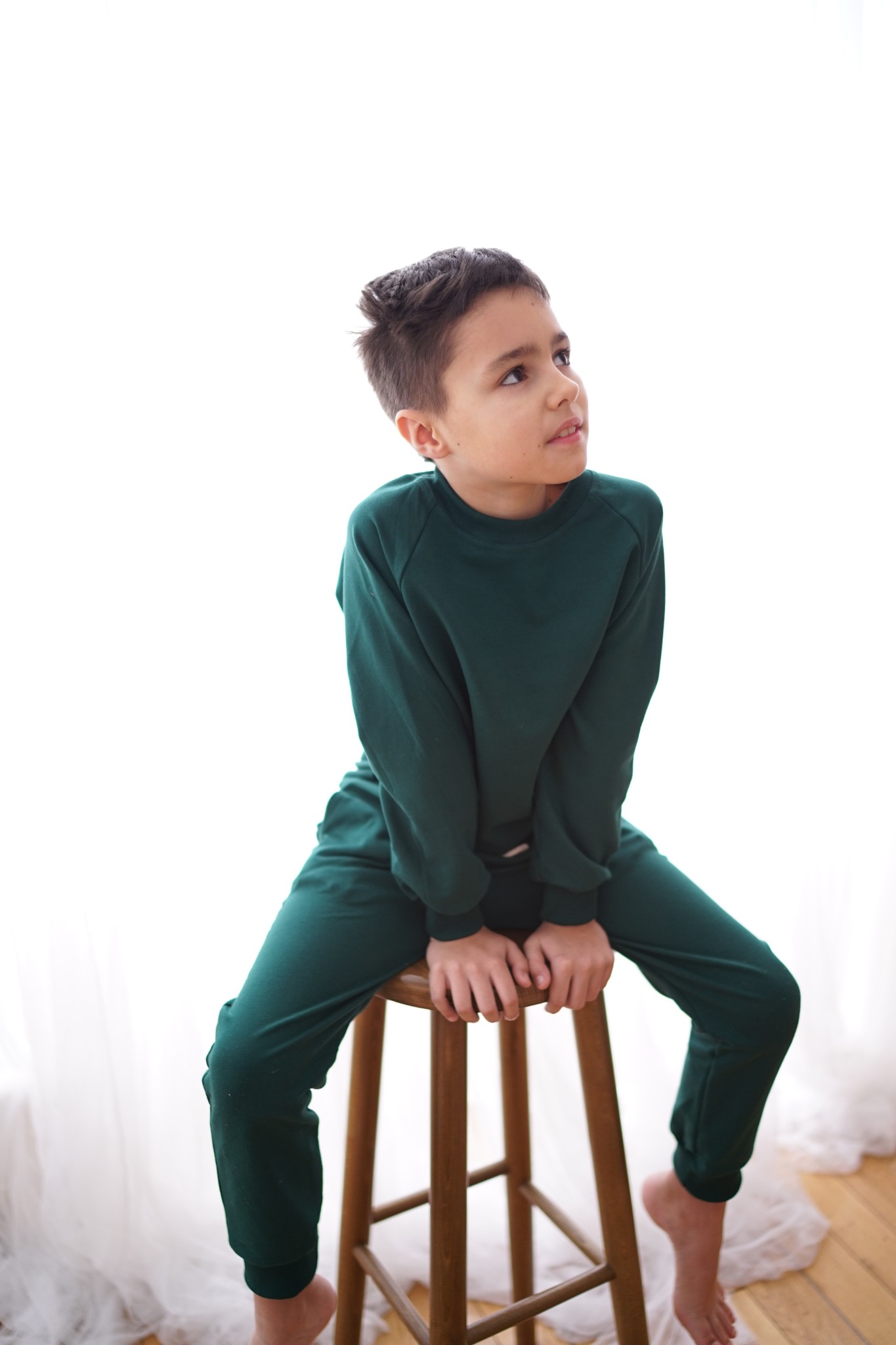 Chłopiec w zielonym dresie siedzi na drewnianym stołku, spoglądając w górę. Jasne, rozproszone światło. Minimalistyczne tło z delikatną firanką.