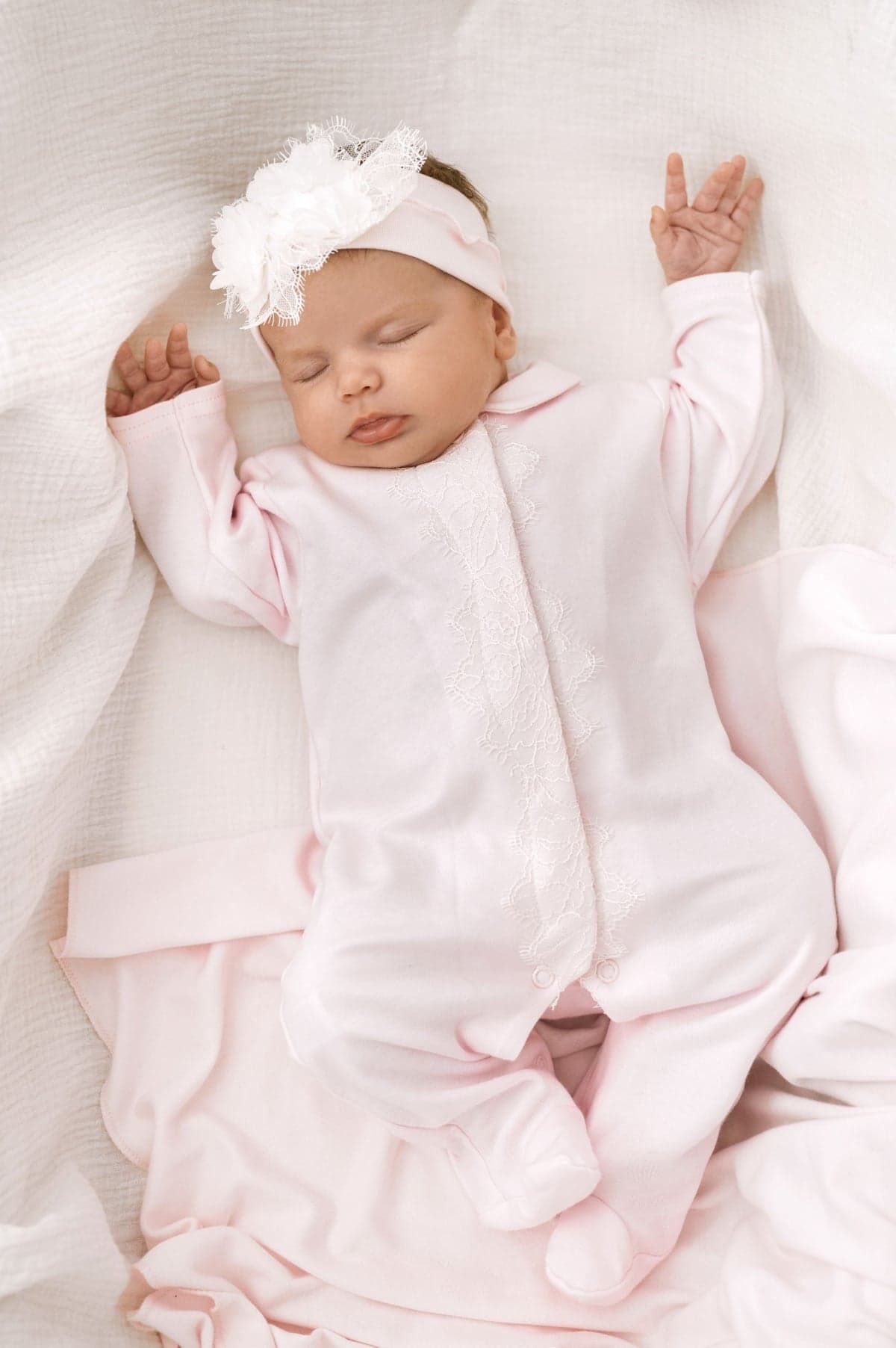 Śpiące niemowlę w różowym pajacyku z koronkowym zdobieniem, leżące na białym, teksturowanym kocyku, z białą opaską z kwiatem na głowie.
