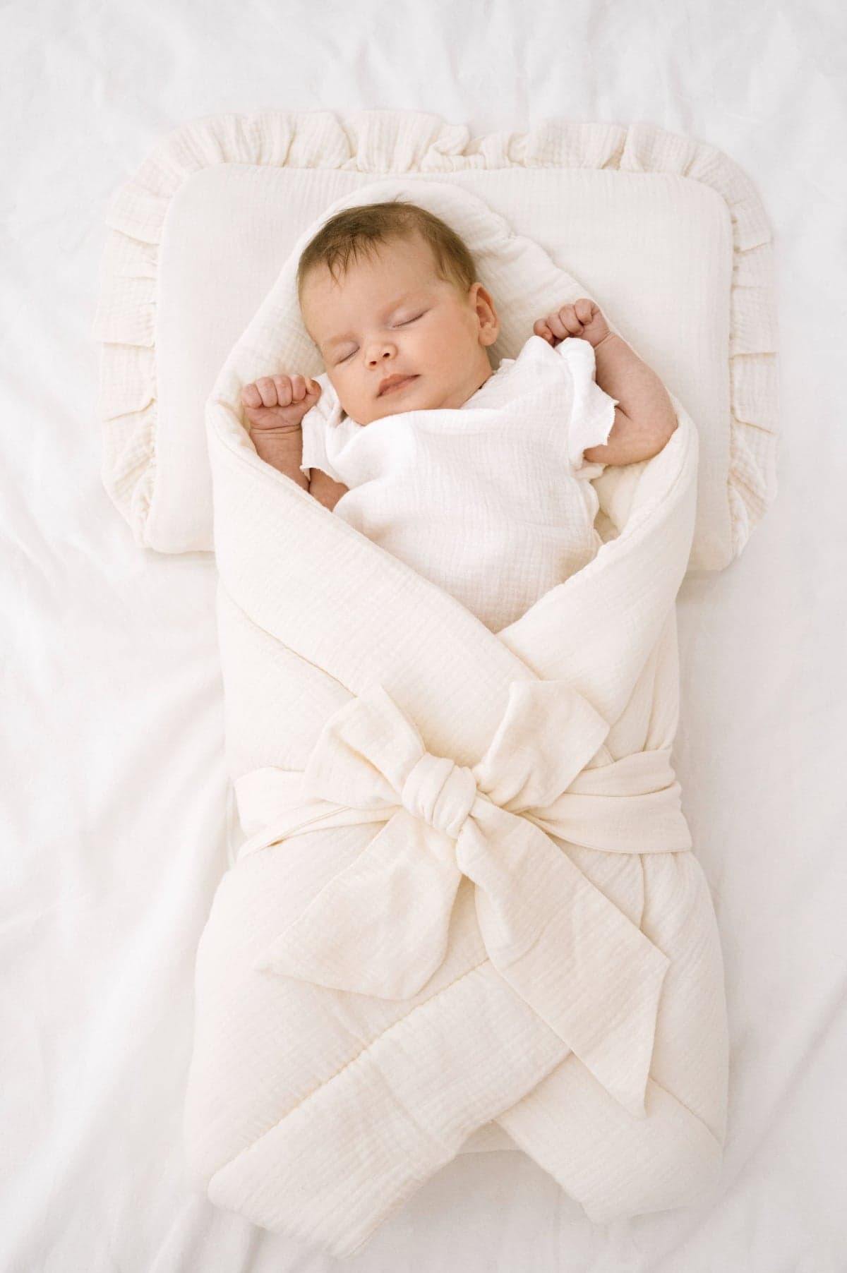 Śpiące niemowlę w białym, muślinowym otulaczu z kokardą, leżące na poduszce z falbanką na białym prześcieradle, ujęcie z góry.