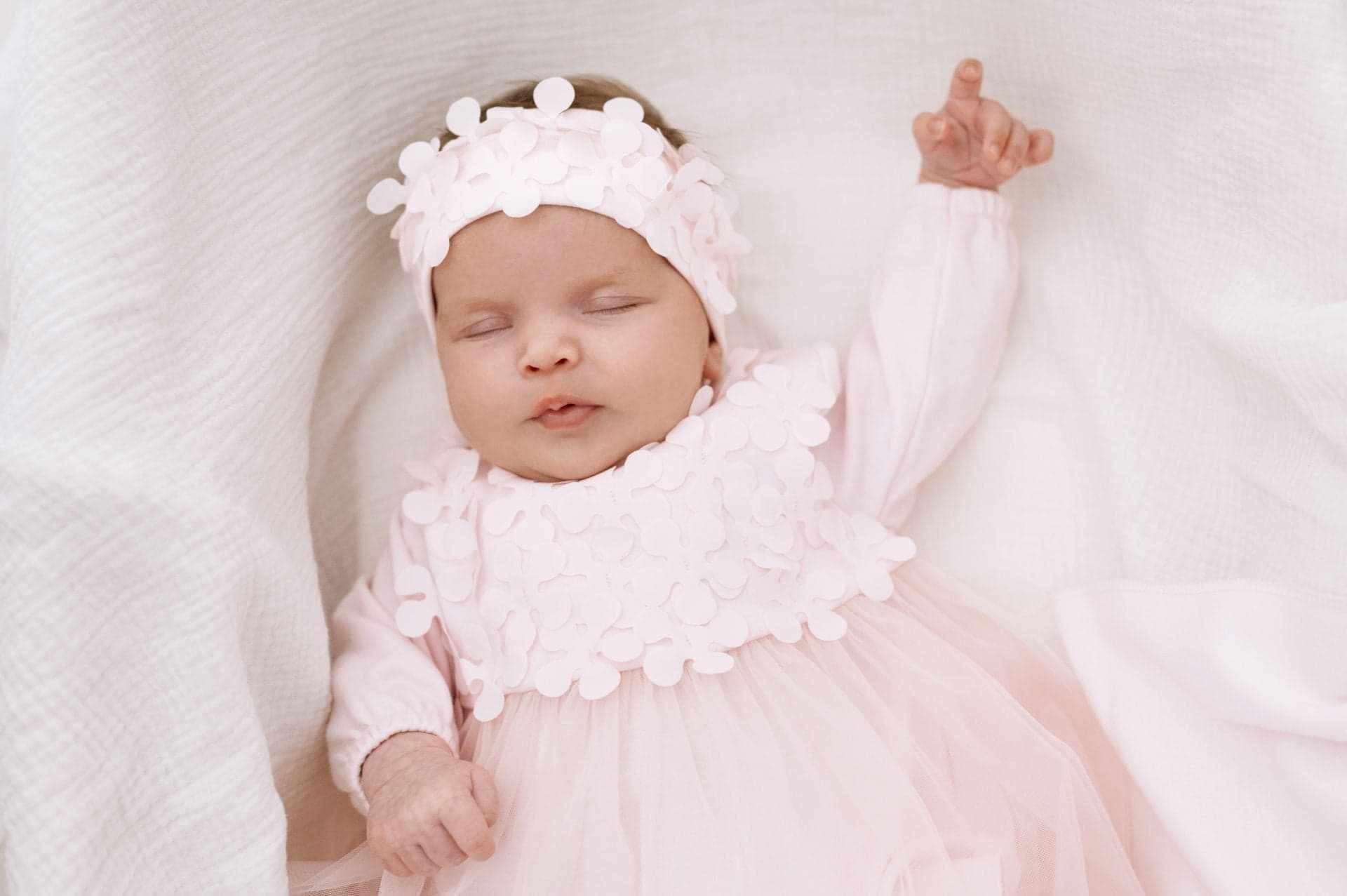 Śpiąca niemowlę w delikatnej, różowej sukience z tiulu i opasce z kwiatkami, leżąca na białej, teksturowanej tkaninie.