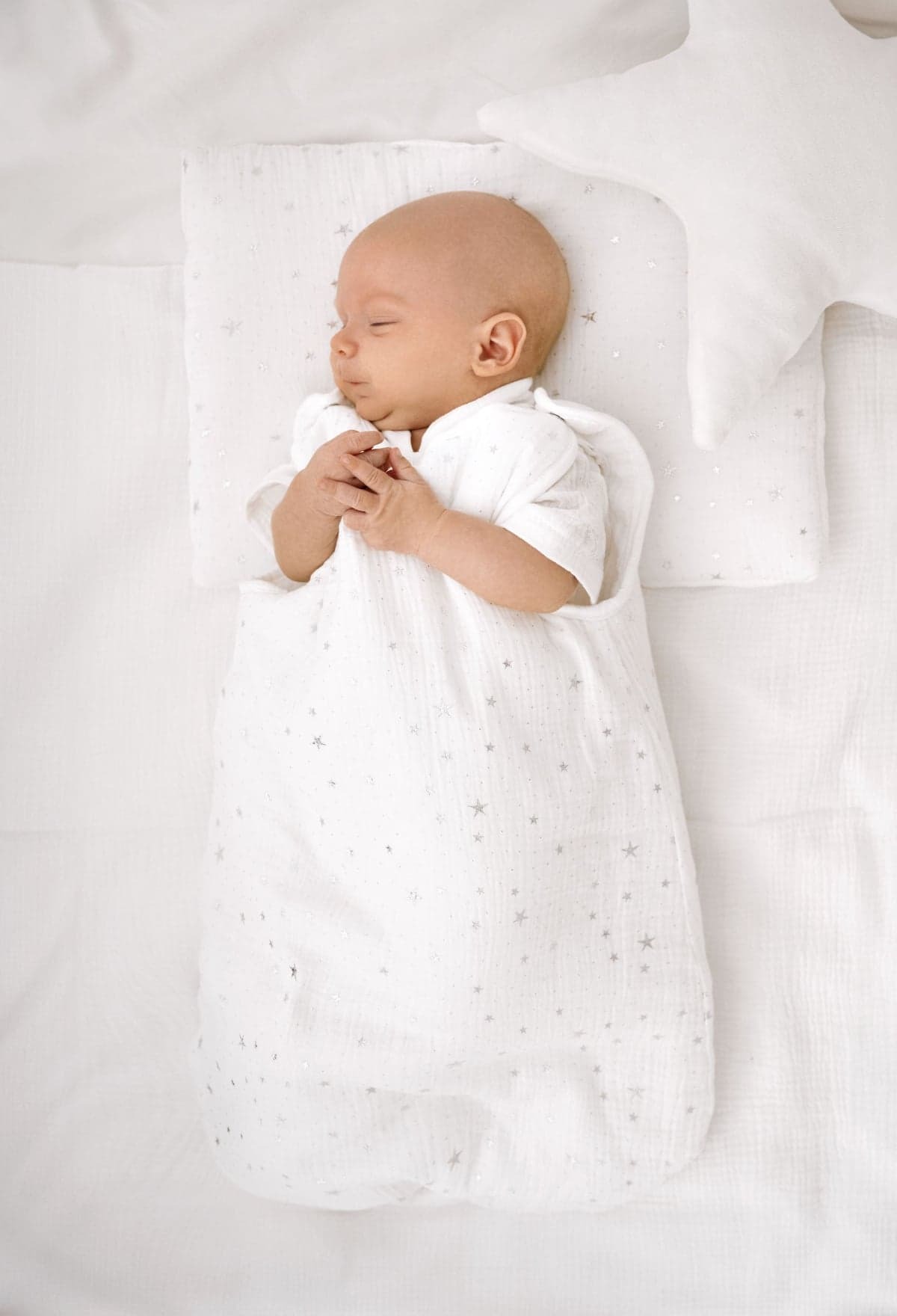 Śpiące niemowlę w białym śpiworku z motywem srebrnych gwiazdek, leżące na białej pościeli obok poduszki w kształcie gwiazdy.