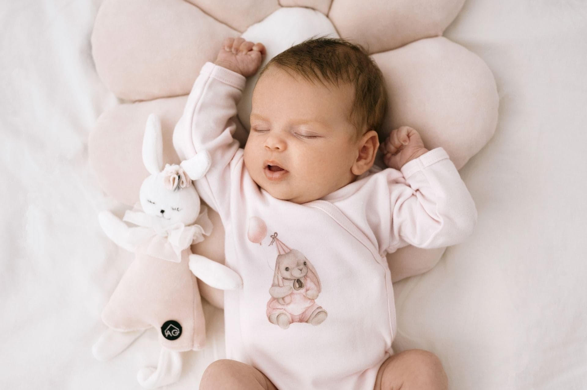 Śpiące niemowlę w różowym body z nadrukiem królika w czapeczce urodzinowej, leżące na poduszce w kształcie kwiatka obok pluszowego króliczka z kokardką.