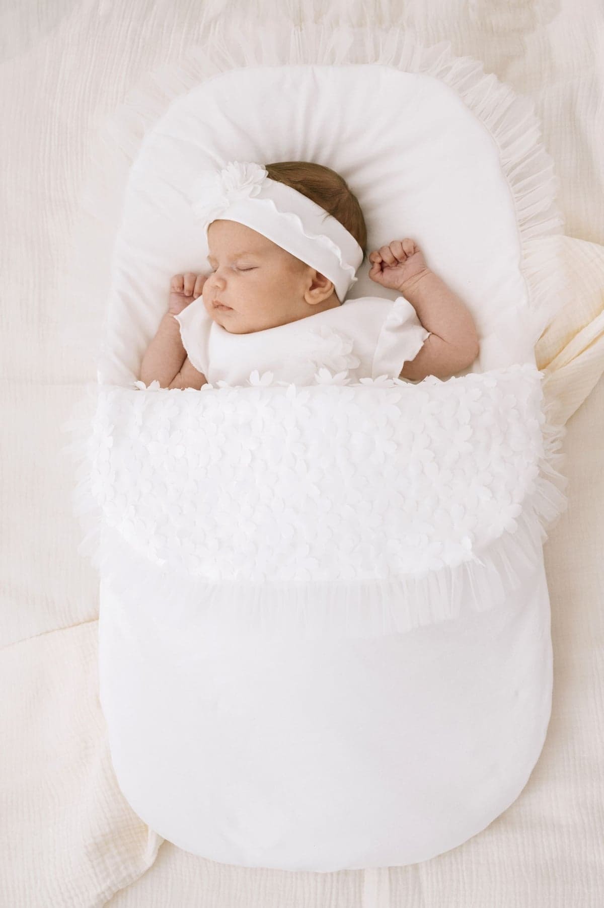 Śpiące niemowlę w białym, eleganckim kokonie z falbankami i kwiatowymi aplikacjami, dopasowana opaska na głowie, delikatne oświetlenie.
