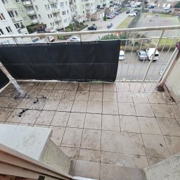 Wiktoria Jackowska - Zabrudzony balkon z widokiem na okoliczne budynki i ulicę, osłonięty czarną siatką, z doniczką z czerwonymi kwiatami przy balustradzie.