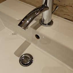 Wiktoria Jackowska - Błyszcząca, chromowana bateria umywalkowa na czystej, białej umywalce z ceramiczną powłoką, widoczne odbicia światła i marmurowe wykończenie ściany w tle.