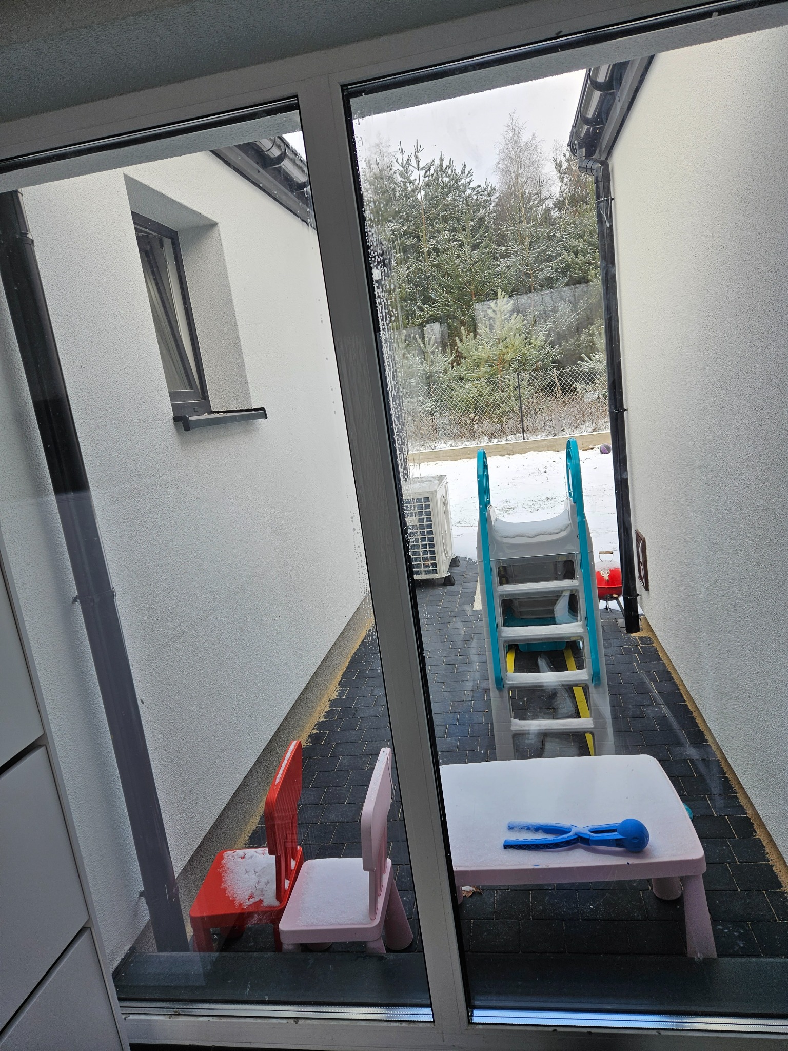 Widok przez okno na ośnieżony plac zabaw z plastikowym stolikiem, krzesełkami i zjeżdżalnią, pomiędzy dwoma budynkami z białego tynku, w tle ośnieżone drzewa.