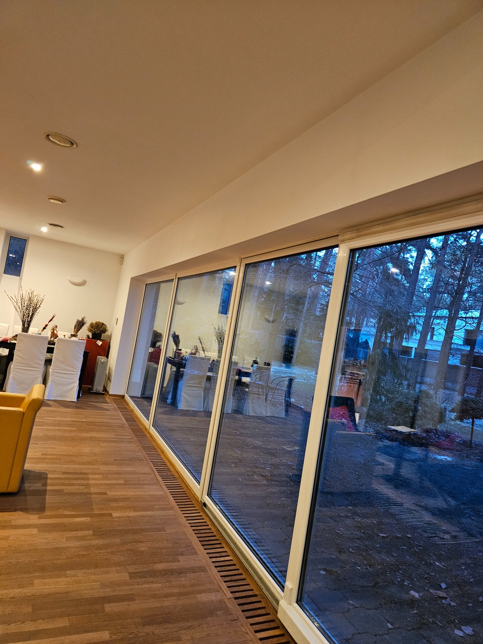 Wnętrze jasnego pomieszczenia z parkietem i dużymi przesuwnymi oknami, przez które widać zimowy krajobraz. Ustawione stoły z białymi pokrowcami na krzesłach, przygotowane do uroczystości. Widoczne...