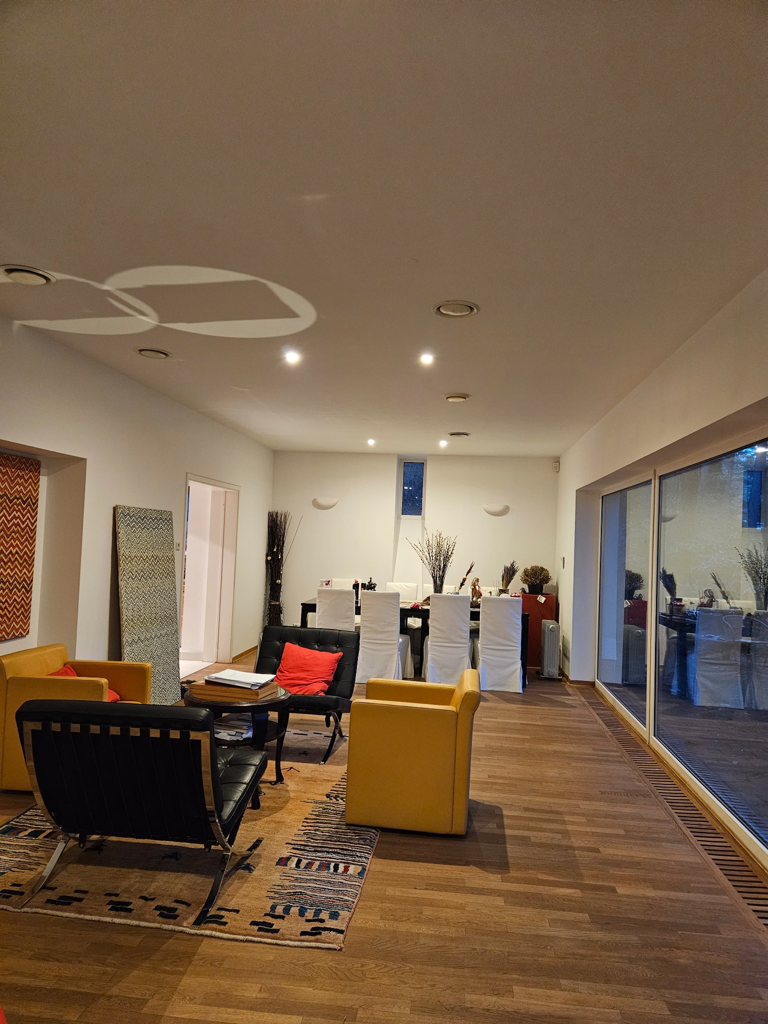 Jasne, przestronne wnętrze domu z drewnianą podłogą, żółtymi i czarnymi fotelami, dywanem w etniczne wzory oraz długim stołem z krzesłami w białych pokrowcach, widoczne przez okno. Na suficie...