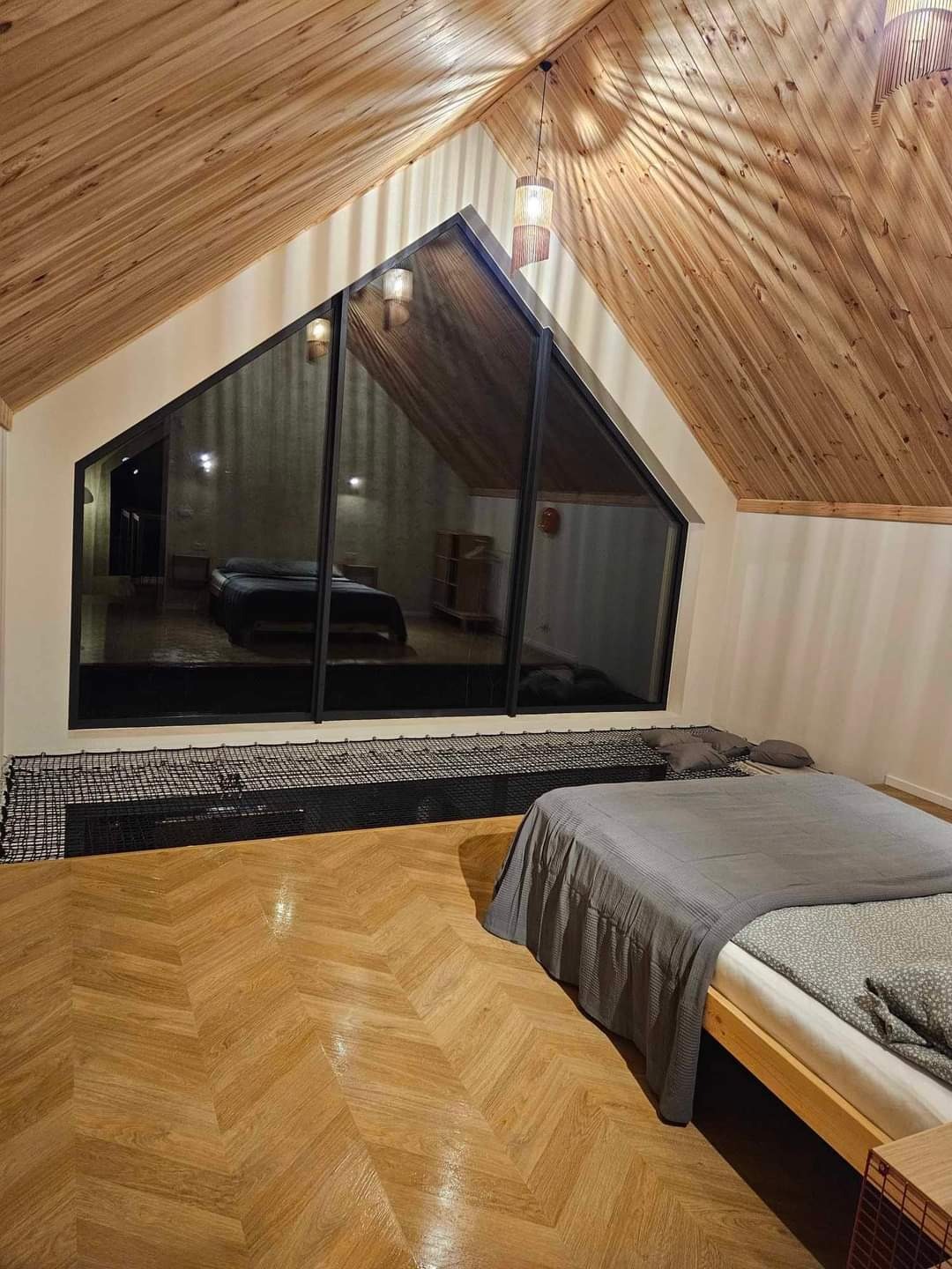 Sypialnia na poddaszu z drewnianym wykończeniem, dużym oknem w kształcie trapezu z widokiem na inny pokój, podłogą w jodełkę i łóżkiem z szarą narzutą.