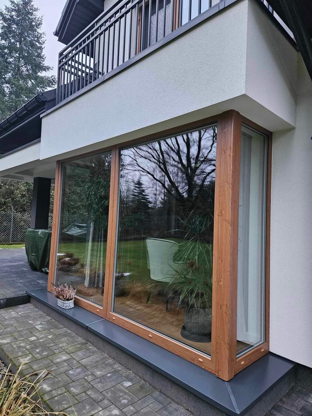 Duże okno narożne z drewnianą ramą, widok z zewnątrz na taras wyłożony kostką, w tle ogród z drzewami.