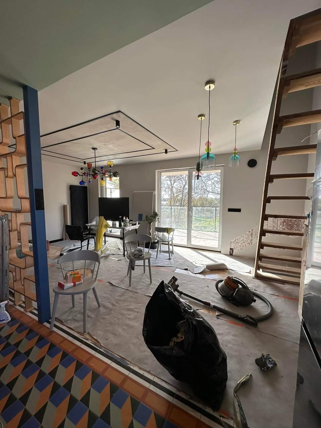 Wnętrze mieszkania w trakcie sprzątania po remoncie, widoczny worek na śmieci, odkurzacz, zabezpieczona podłoga i meble, w tle kolorowe lampy i schody.