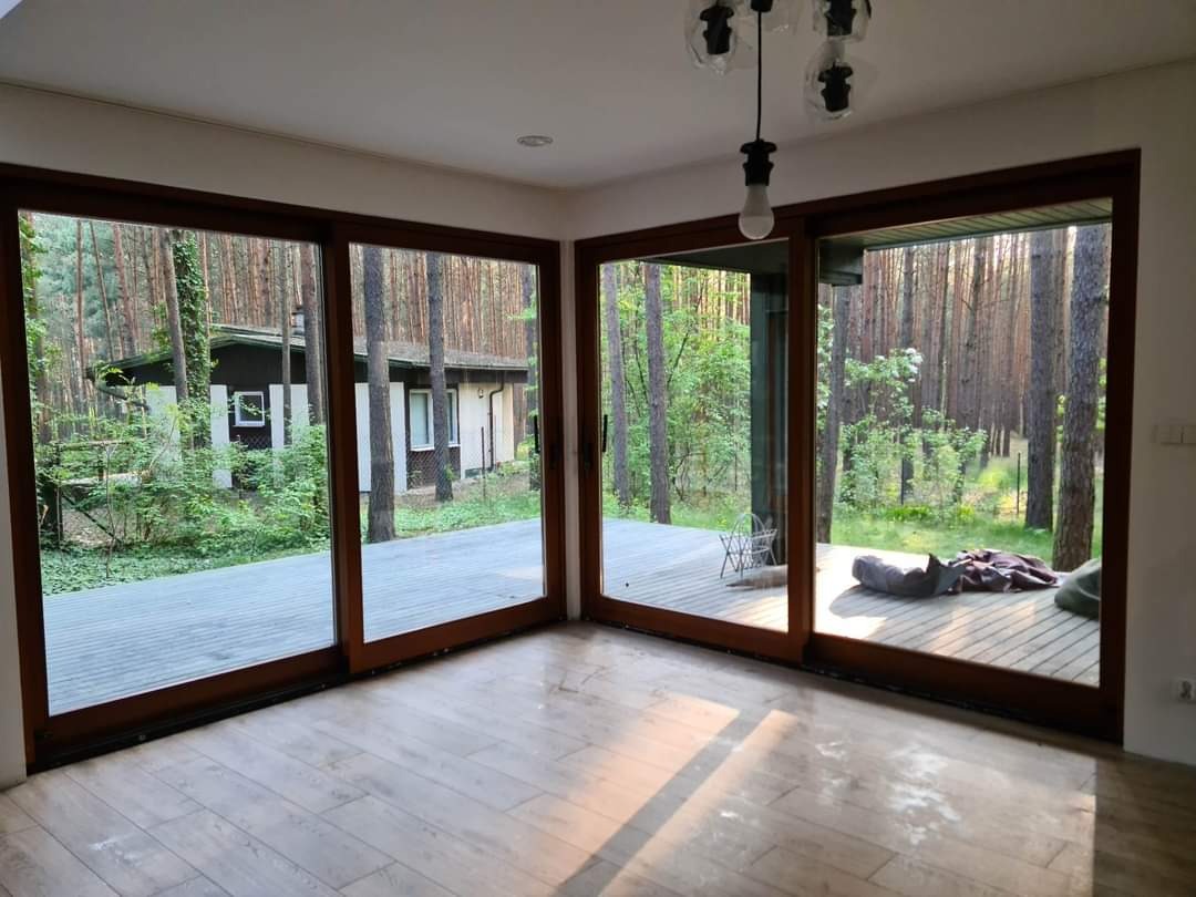 Puste pomieszczenie z dużymi przesuwnymi oknami z brązowymi ramami, widok na zalesiony ogród z drewnianym tarasem, podłoga wyłożona panelami imitującymi drewno, surowa żarówka zwisająca z sufitu.