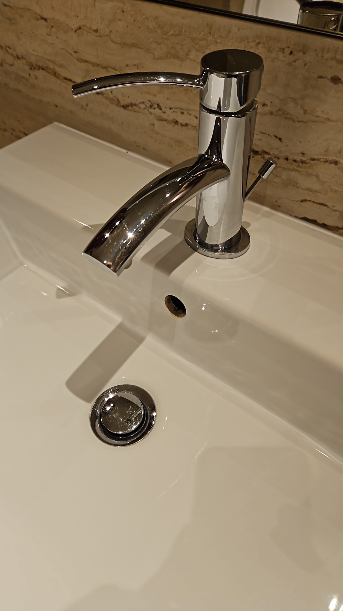 Błyszcząca, chromowana bateria umywalkowa na czystej, białej umywalce z ceramiczną powłoką, widoczne odbicia światła i marmurowe wykończenie ściany w tle.