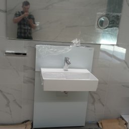 Błażej Grzesiak - Biała umywalka z chromowaną baterią, zabezpieczona folią, zamontowana na białej szafce, na tle marmurowych płytek, widoczne odbicie osoby w lustrze i prowizoryczne zabezpieczenie podłogi.