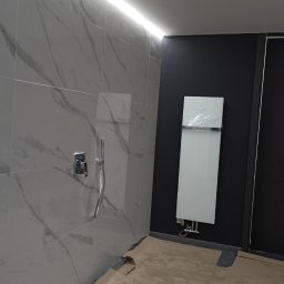 Błażej Grzesiak - Łazienka w trakcie remontu: marmurowe płytki na ścianie, zamontowana bateria prysznicowa, grzejnik drabinkowy w folii ochronnej na czarnej ścianie, podłoga zabezpieczona kartonem, oświetlenie LED...