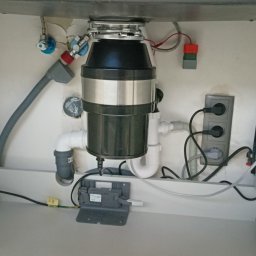 Błażej Grzesiak - Wnętrze szafki pod zlewem z zamontowanym rozdrabniaczem odpadów spożywczych, widoczne podłączenia hydrauliczne, elektryczne i gniazdko.