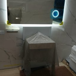 Błażej Grzesiak - Podświetlane lustro łazienkowe z wyświetlaczem czasu, zabezpieczone żółtymi narożnikami ochronnymi, umywalka zakryta folią ochronną w trakcie wykańczania łazienki.