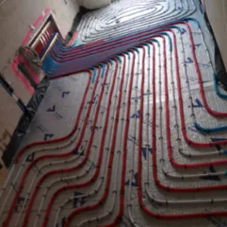 Rozłożona instalacja ogrzewania podłogowego z czerwonych, białych i niebieskich rur na srebrnej folii izolacyjnej w surowym wnętrzu z widocznym oknem dachowym i rozdzielaczem na ścianie.