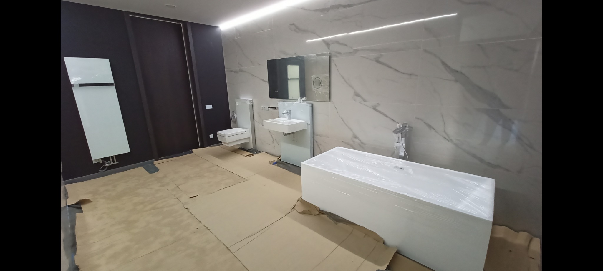 Wnętrze łazienki w trakcie prac wykończeniowych, z zabezpieczoną folią wanną, umywalką i toaletą na tle marmurowych płytek ściennych i brązowych drzwi.