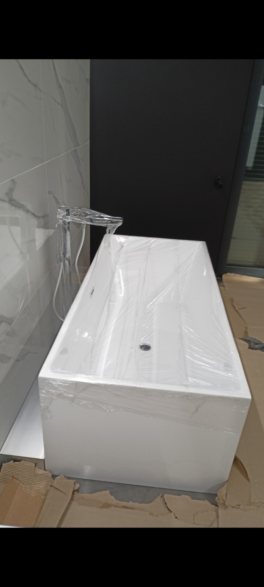 Nowa, prostokątna wanna z baterią, owinięta folią ochronną, stojąca na kartonach podczas montażu w łazience z marmurowymi płytkami.