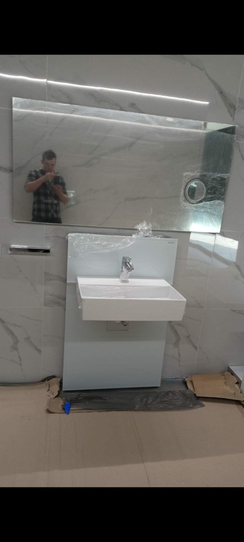 Biała umywalka z chromowaną baterią, zabezpieczona folią, zamontowana na białej szafce, na tle marmurowych płytek, widoczne odbicie osoby w lustrze i prowizoryczne zabezpieczenie podłogi.