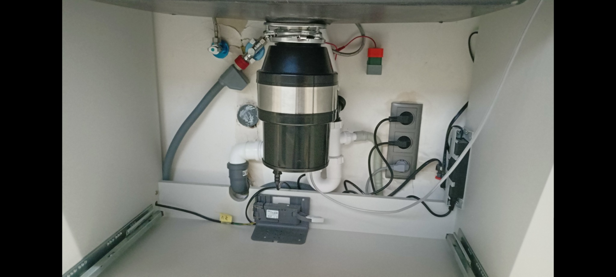 Wnętrze szafki pod zlewem z zamontowanym rozdrabniaczem odpadów spożywczych, widoczne podłączenia hydrauliczne, elektryczne i gniazdko.