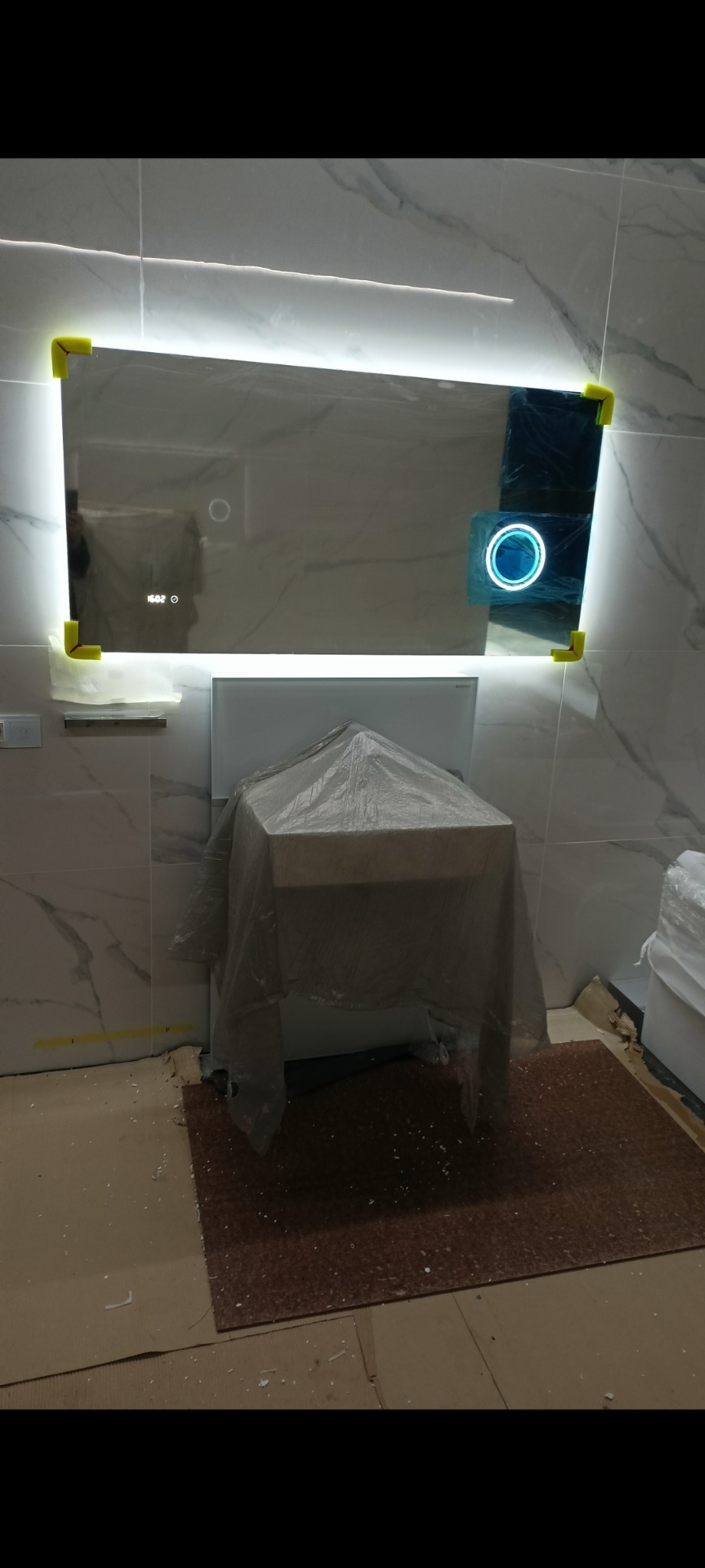 Podświetlane lustro łazienkowe z wyświetlaczem czasu, zabezpieczone żółtymi narożnikami ochronnymi, umywalka zakryta folią ochronną w trakcie wykańczania łazienki.