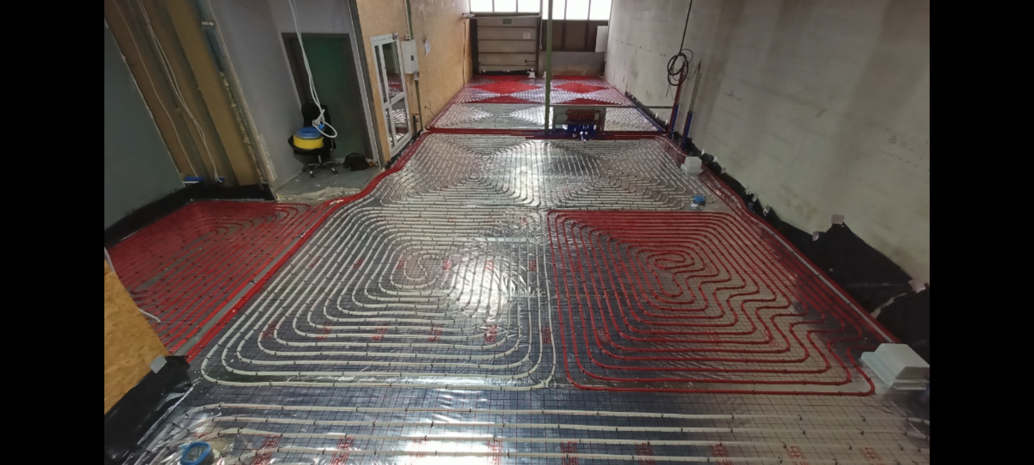 Rozłożone rury ogrzewania podłogowego w dużym pomieszczeniu, czerwone i białe rury ułożone w pętle na folii z siatką, widok z góry na etapie instalacji, surowy stan wykończenia.