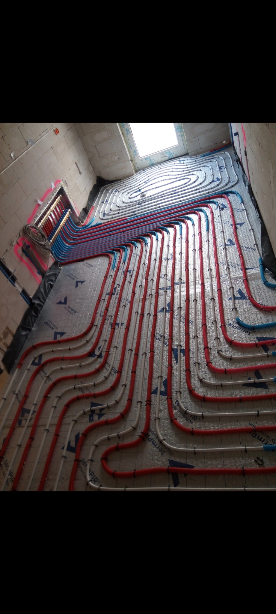 Rozłożona instalacja ogrzewania podłogowego z czerwonych, białych i niebieskich rur na srebrnej folii izolacyjnej w surowym wnętrzu z widocznym oknem dachowym i rozdzielaczem na ścianie.