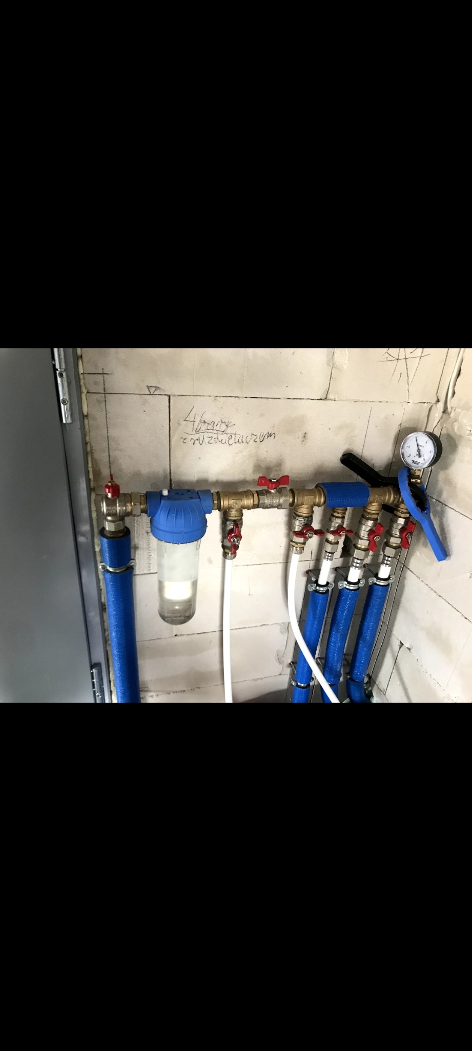 Rozdzielacz hydrauliczny z zaworami, filtrem i manometrem zamontowany na ścianie z surowego betonu z widocznymi śladami ołówka.