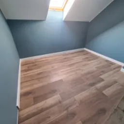 Poddasze po remoncie: świeżo pomalowane na błękitno ściany, jasne listwy przypodłogowe i panele podłogowe imitujące drewno, widoczne okno dachowe.