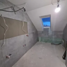 Poddasze w trakcie wykańczania: szare płytki na ścianach, widoczne instalacje elektryczne i hydrauliczne, okno dachowe z widokiem na niebo.
