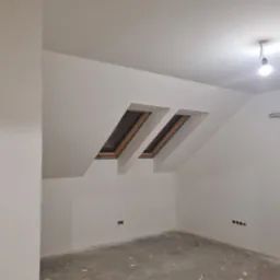 Poddasze po malowaniu na biało, z dwoma oknami dachowymi i widocznymi gniazdkami elektrycznymi, surowa żarówka i kabel na ścianie.