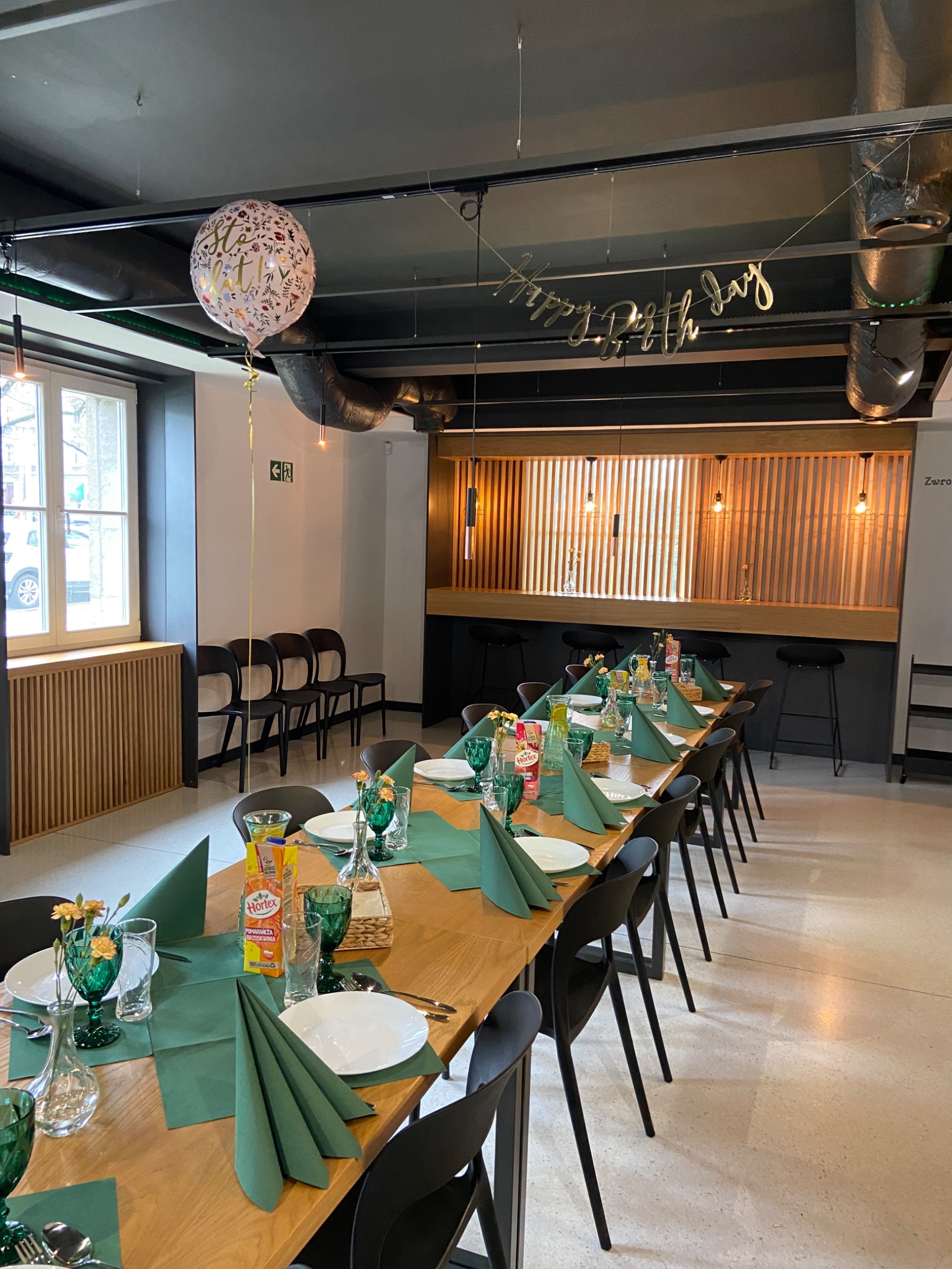 Uroczysty stół urodzinowy z zielonymi akcentami: serwetki, szklanki i małe bukiety kwiatów. Balon z napisem 'Sto lat' i złoty napis 'Happy Birthday' zawieszone pod sufitem. Widoczne krzesła...