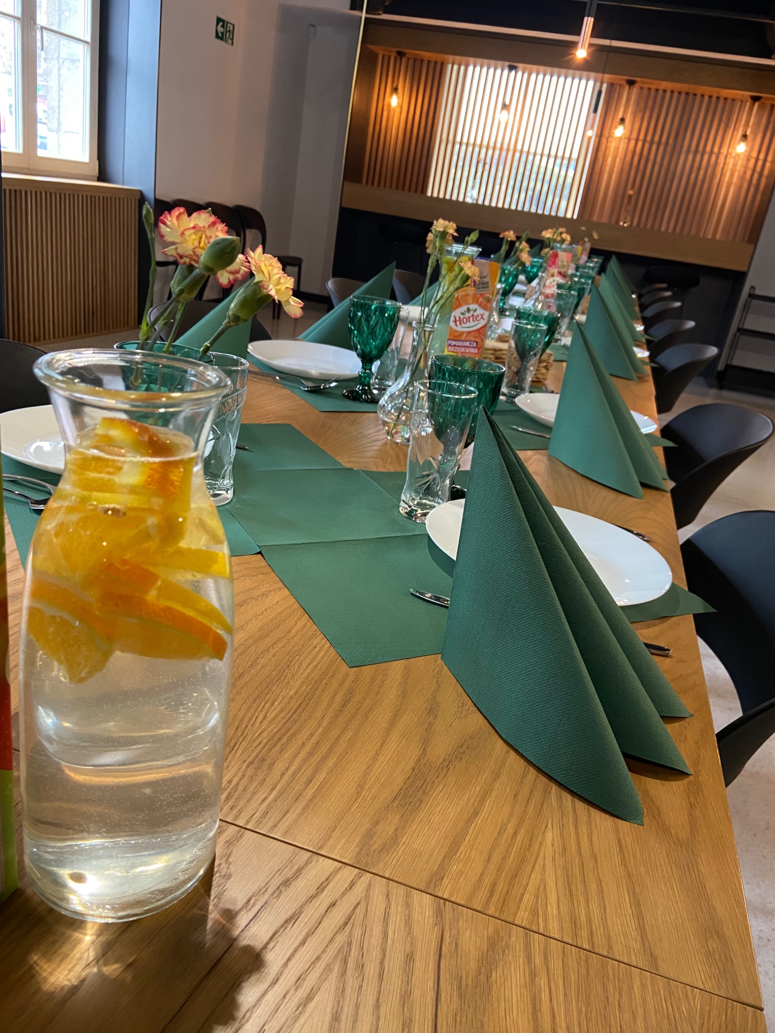 Długi stół z drewnianym blatem, nakryty zielonymi bieżnikami i serwetkami w kształcie stożków, białymi talerzami, zielonymi szklankami, dzbankiem z wodą i pomarańczami oraz kwiatami w wazonach...