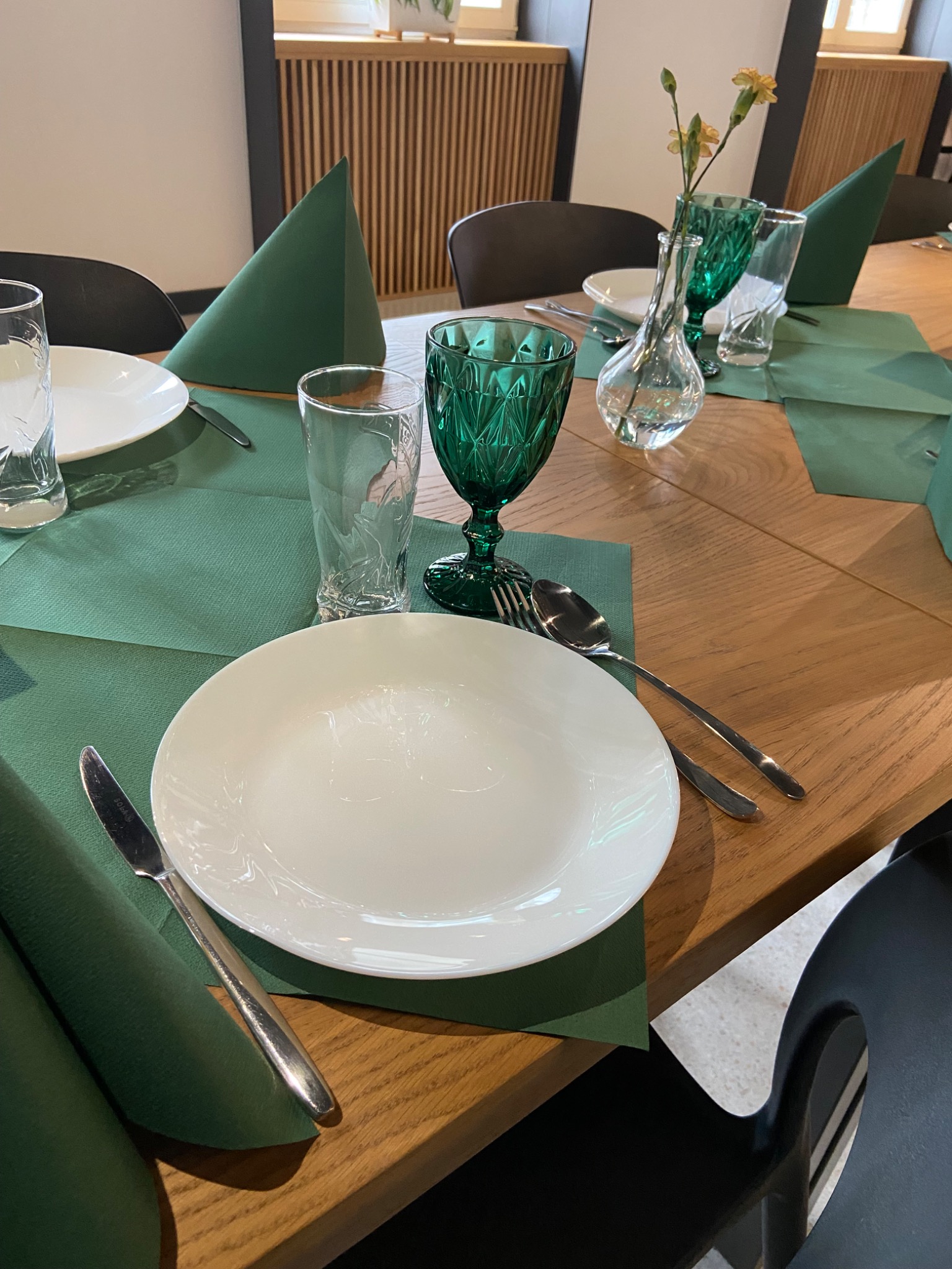 Elegancko nakryty stół: białe talerze, zielone serwetki, kryształowe kieliszki i wazonik z drobnymi kwiatami, drewniany blat stołu.