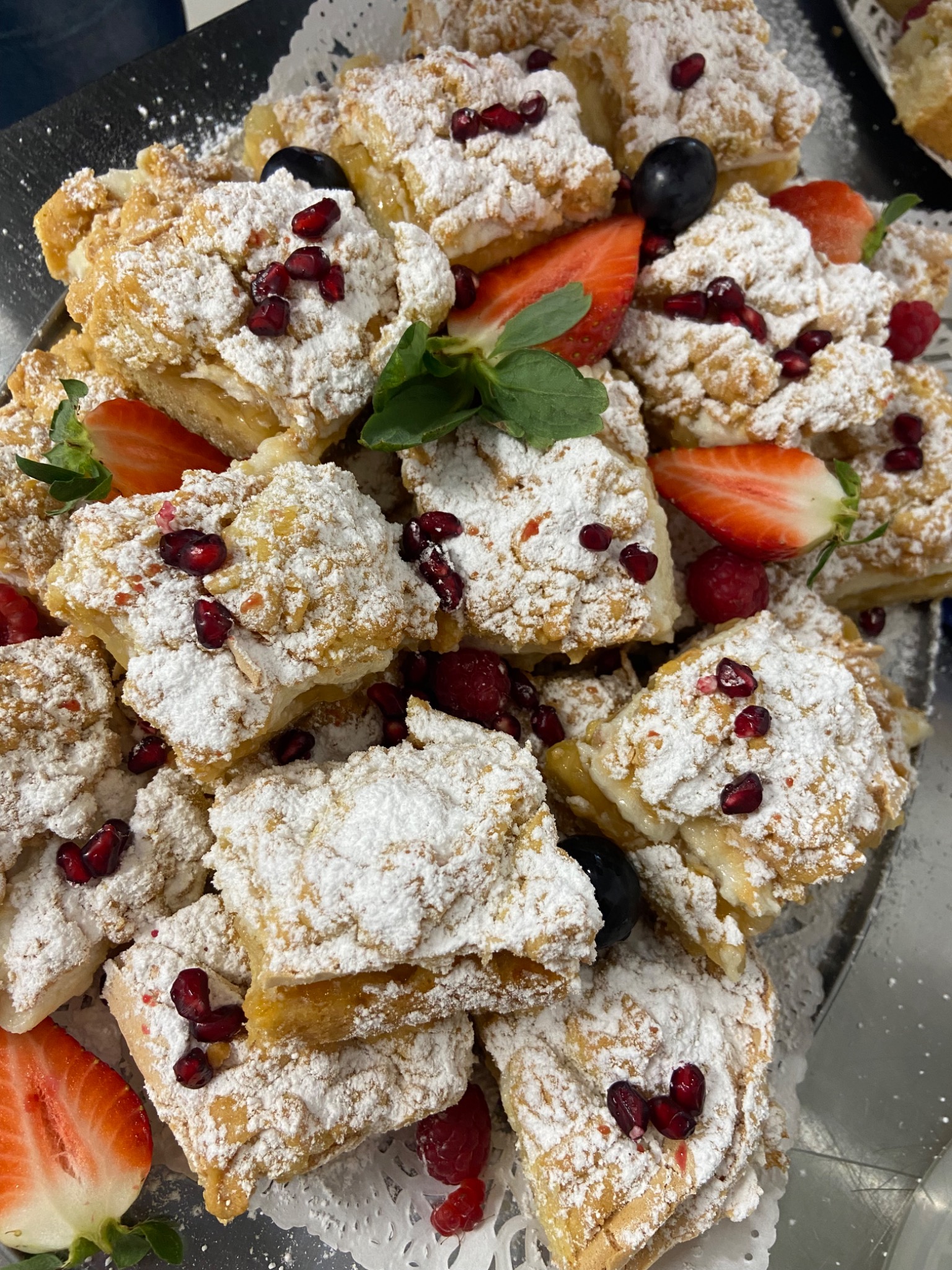 Kwadratowe kawałki ciasta posypane cukrem pudrem i udekorowane owocami granatu, truskawkami, malinami i listkiem mięty na srebrnej tacy.