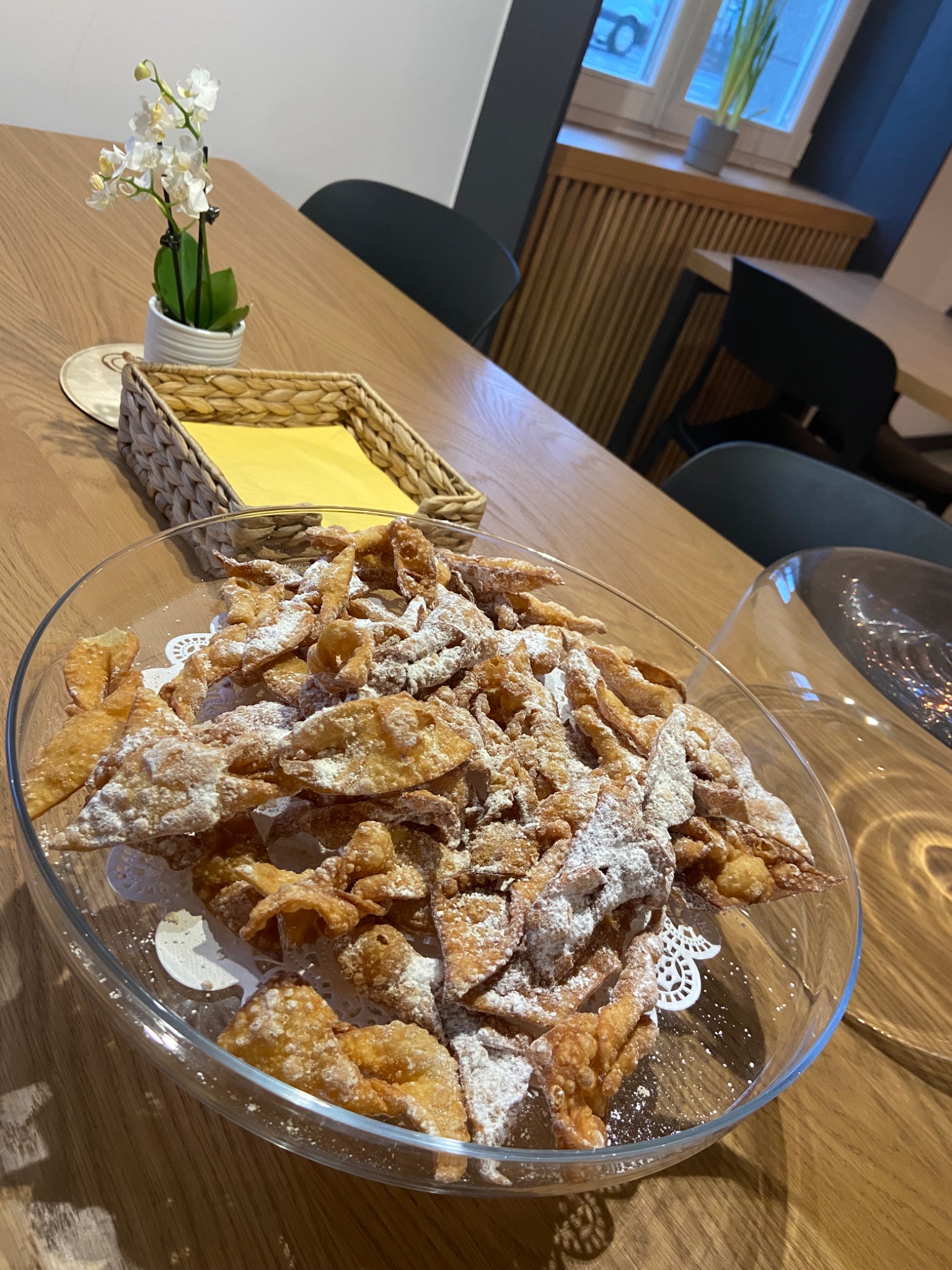 Szklana misa wypełniona chrustem posypanym cukrem pudrem, umieszczona na drewnianym stole obok dekoracyjnego koszyka z serwetkami i małej doniczki z białymi kwiatami.