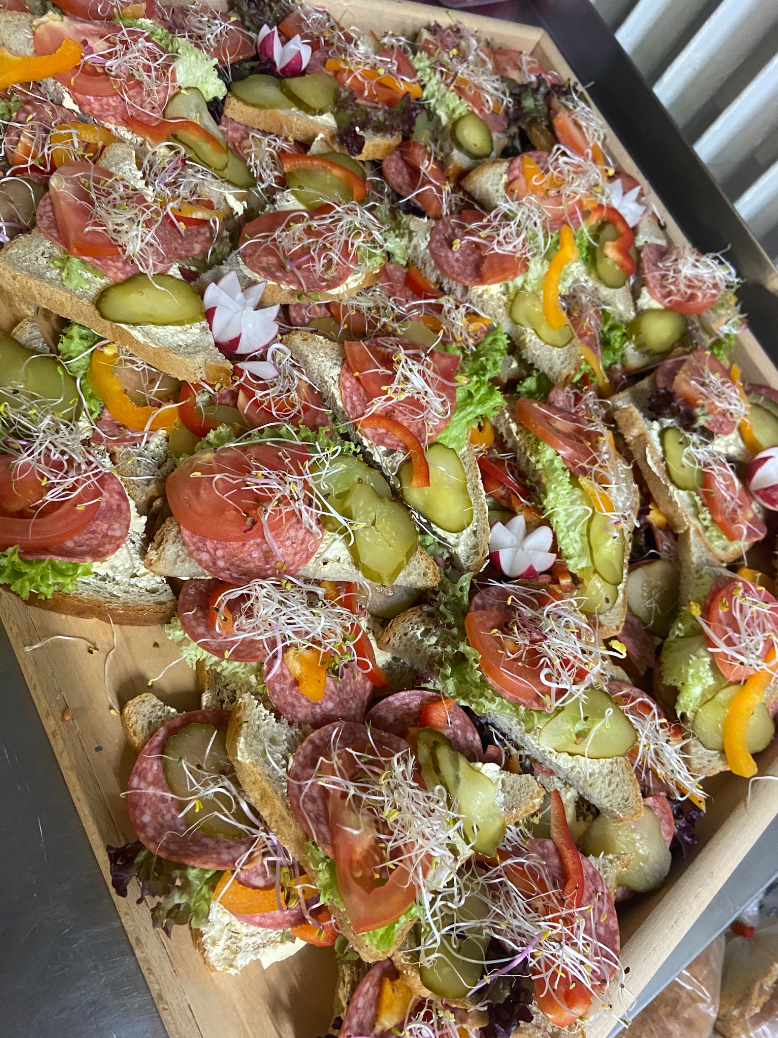 Duża drewniana taca wypełniona kanapkami z salami, pomidorem, ogórkiem kiszonym, papryką, rzodkiewką i kiełkami, ułożonymi blisko siebie.