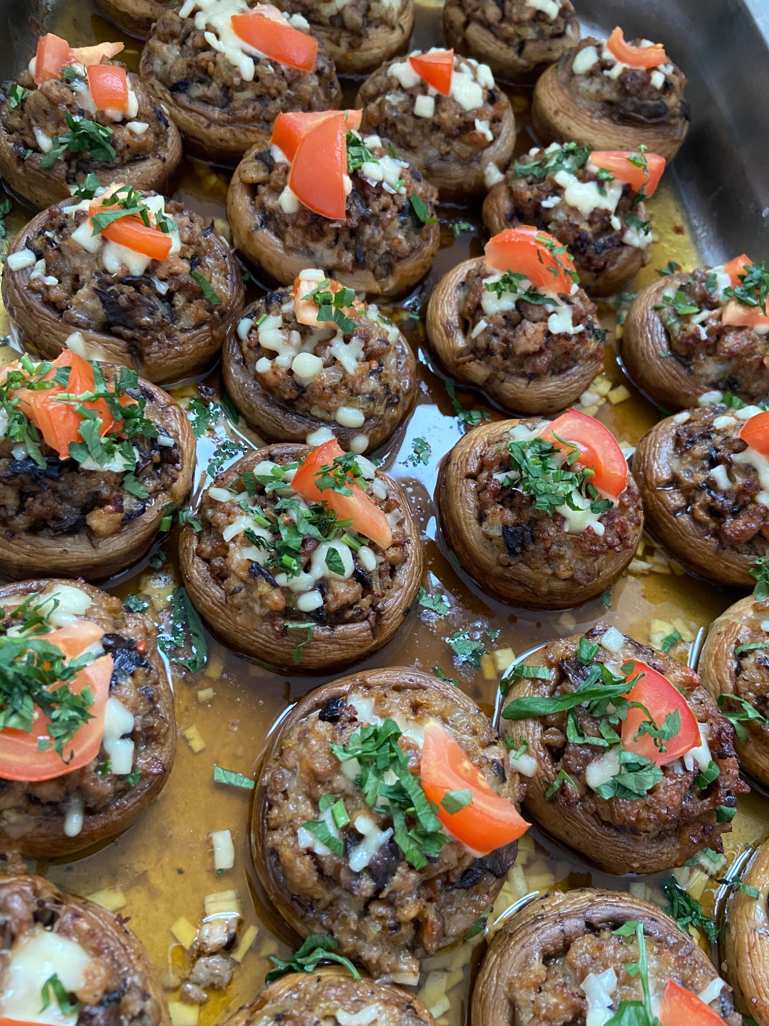 Taca z pieczarkami faszerowanymi mięsem, serem i pomidorami, udekorowanymi świeżą zieloną pietruszką, zanurzonymi w sosie.