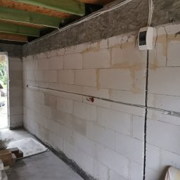 Alex Elektryk Aleksander Łusiarczyk - Wewnętrzna ściana budynku w trakcie prac instalacyjnych, widoczne kable elektryczne prowadzone po ścianie i pod sufitem oraz skrzynka rozdzielcza zamontowana na ścianie.