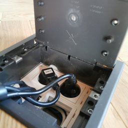 Alex Elektryk Aleksander Łusiarczyk - Otwarta podłogowa puszka elektryczna z dwoma gniazdami, wpięte kable zasilające, widoczne elementy montażowe i przewody wewnątrz.