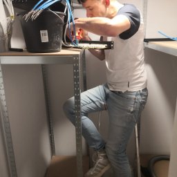 Alex Elektryk Aleksander Łusiarczyk - Młody mężczyzna na drabinie, podłącza kable telekomunikacyjne do panelu krosowniczego, używając zaciskarki. Wiązka kabli w czarnym wiadrze obok.