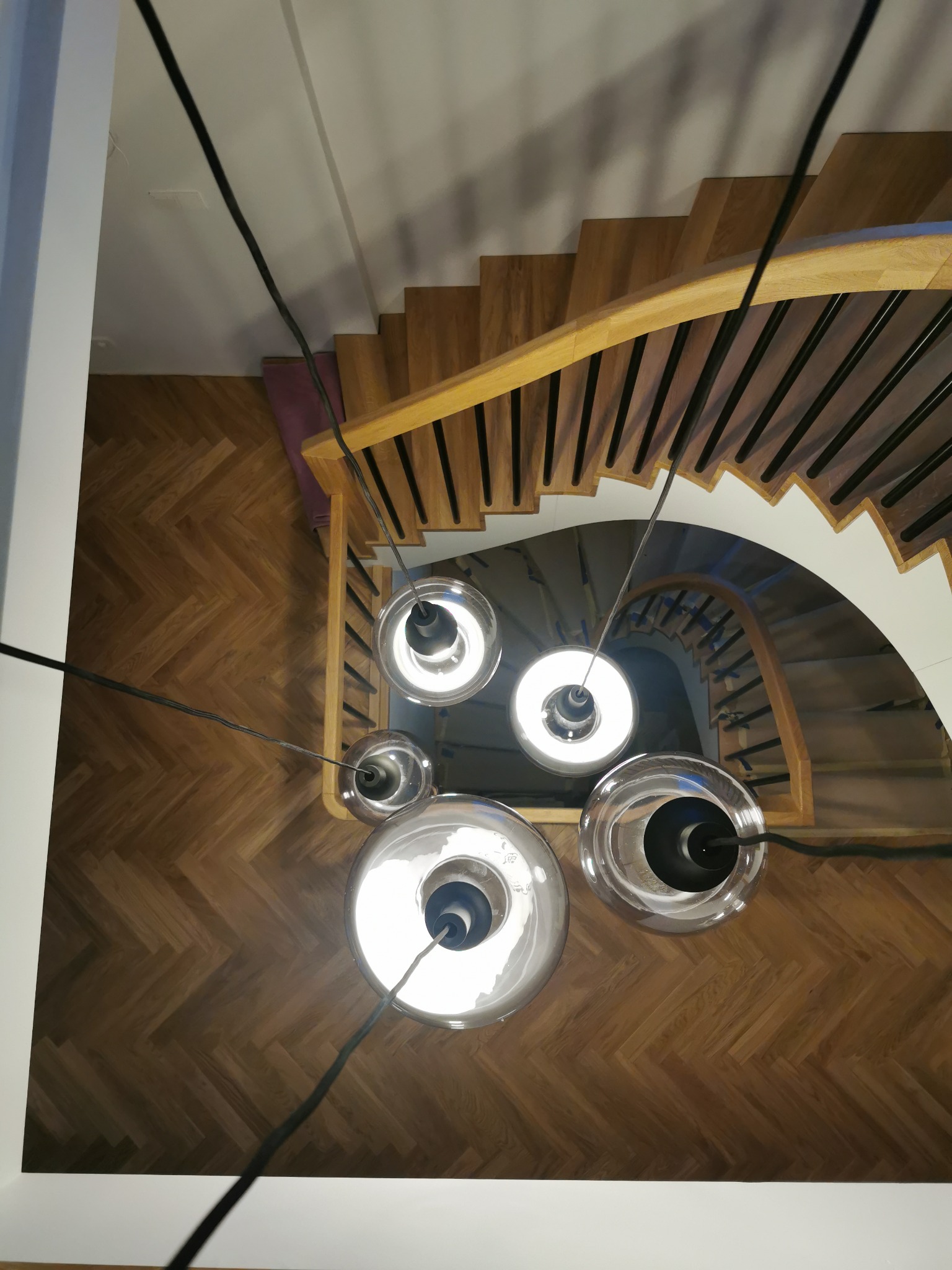 Widok z dołu na spiralne schody z drewnianymi stopniami i czarnymi balustradami, oświetlone przez wiszące lampy z przezroczystymi kloszami i czarnymi przewodami, na tle parkietu ułożonego w jodełkę.
